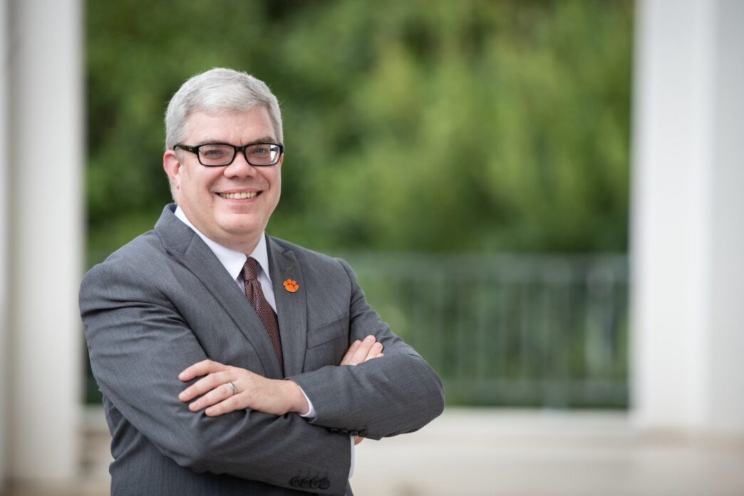 Dean Christopher Cox, in a grey suit, stands smiling with arms folded on library bridge.