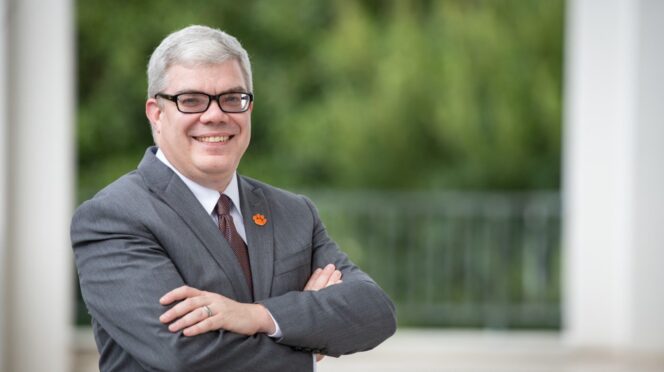 Dean Christopher Cox, in a grey suit, stands smiling with arms folded on library bridge.