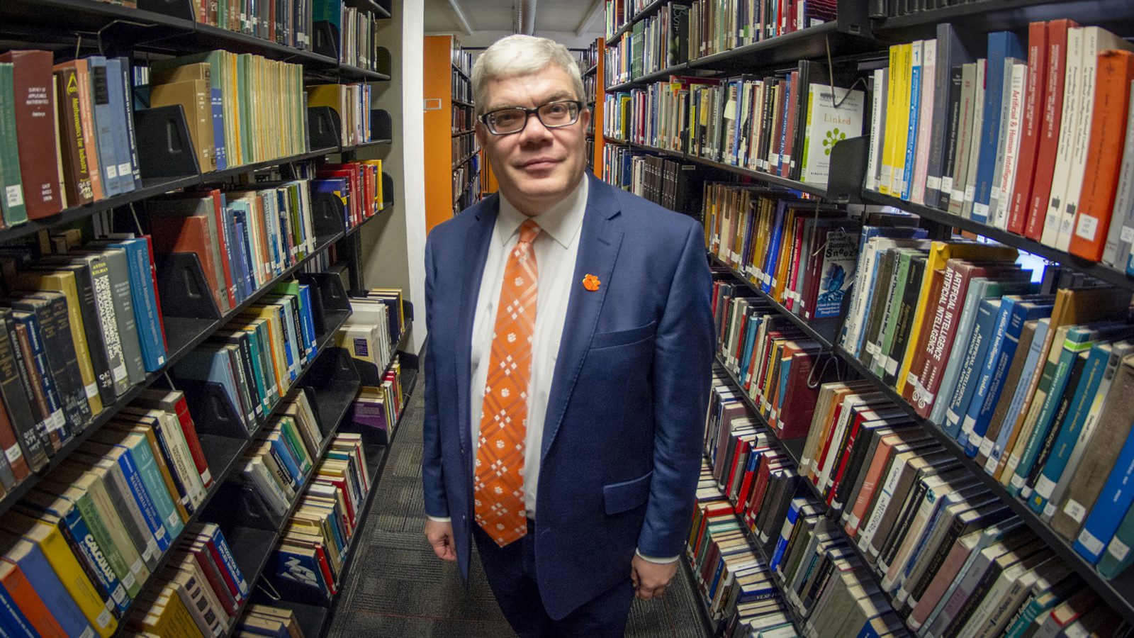 Dean Cox, in a dark blue suit with orange tie, stands between two bookshelves full of books.