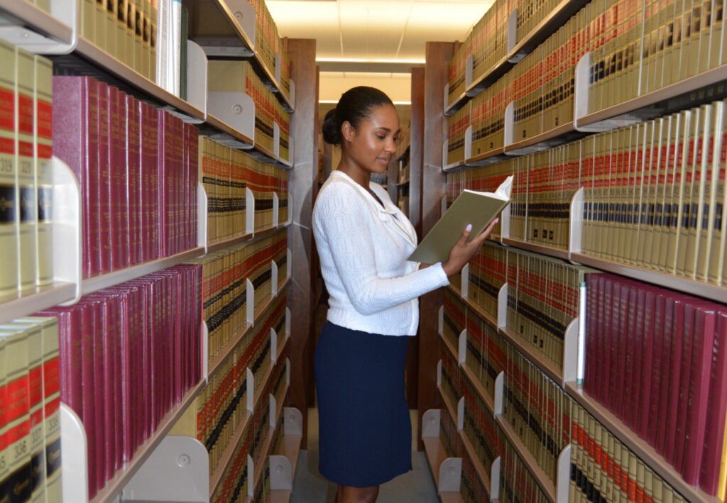 A student stands in the middle of a law library aisle, facing the left of the aisle with a case book in her hand.
