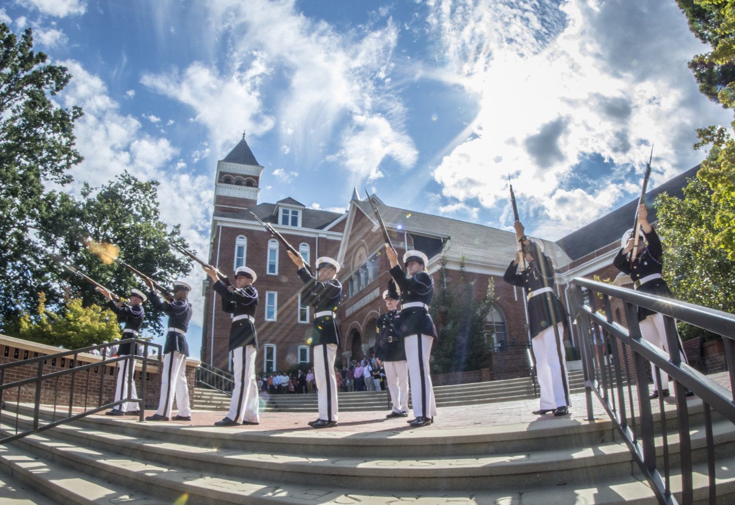 Five Army cadets in dress blue uniforms stand in formation in front of Tillman Hall, pointing rifles up towards the upper left corner of the frame and firing.