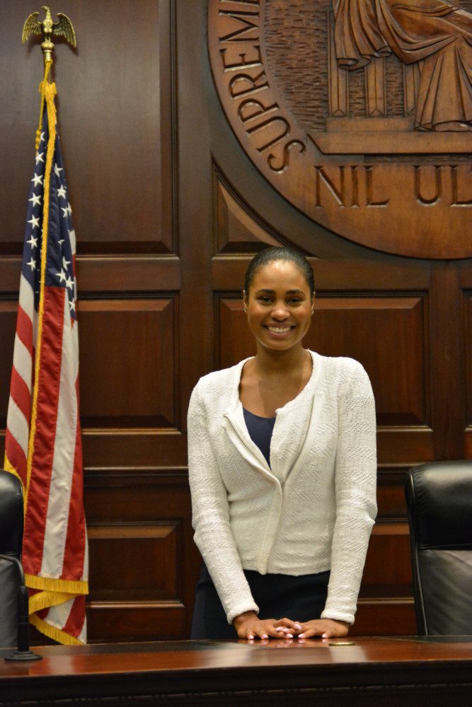 A student leans slightly over a wooden desk. An American flag stands on her right. A wooden seal for the state Supreme Court is hanging behind her.