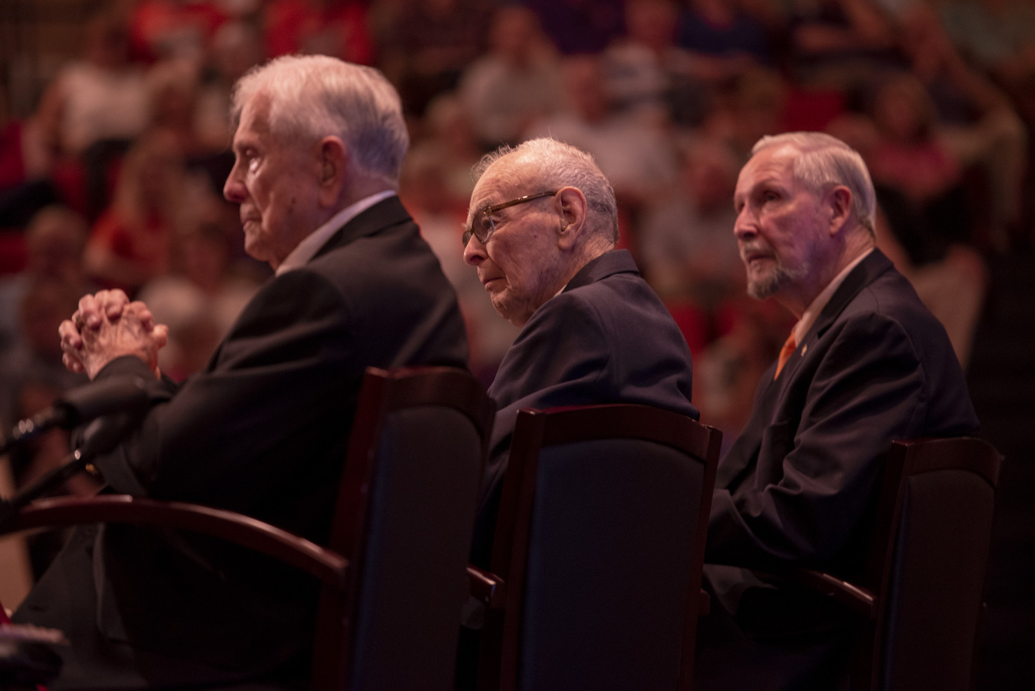 Profile shot of three men in suits with audience in the background