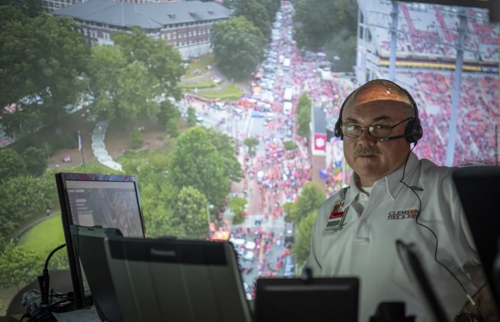 A man studies a computer while the entire wall behind him shows an overhead video of the crowds gathering around Memorial Stadium.