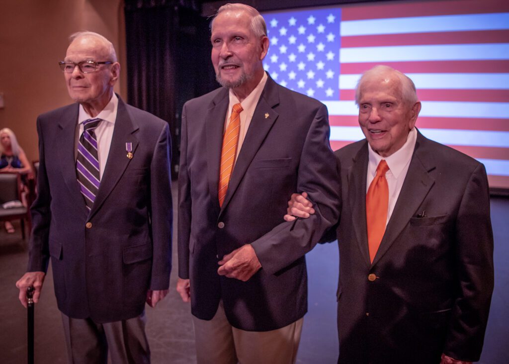 Three men in suits stand in front of an American flag projected on a screen behind them