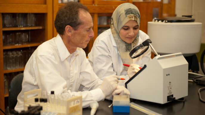Professor Paul Dawson is seated at a lab table looking at a microscope with one of his graduate students