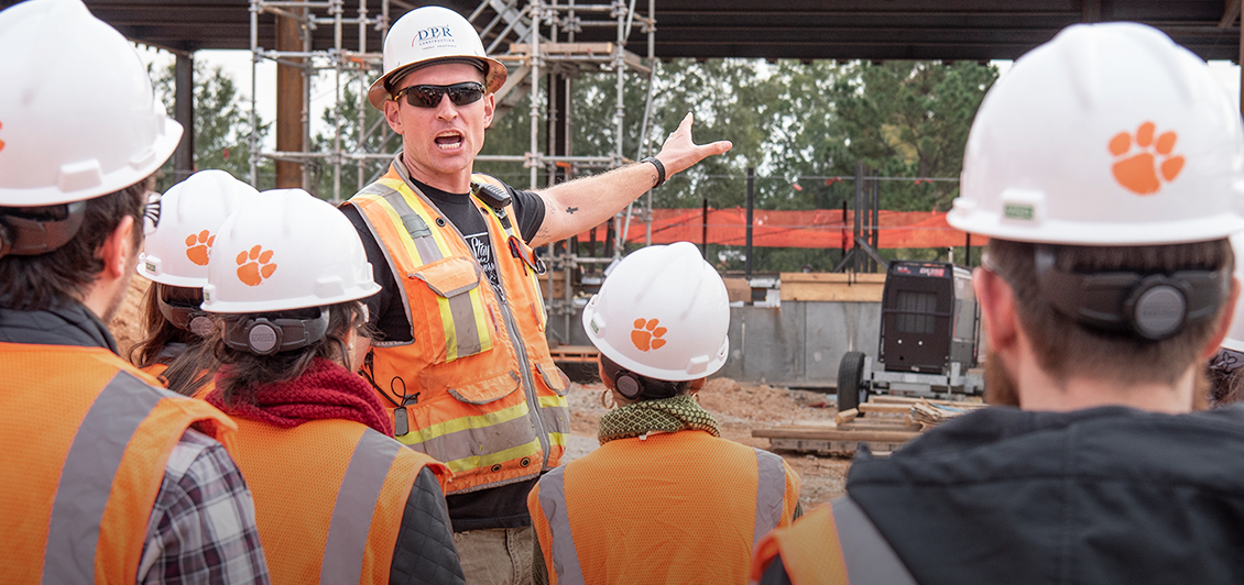 A DPR representative is seen pointing to the construction behind him as a group of students in white hardhats with an orange Tiger Paw are seen in the foreground.