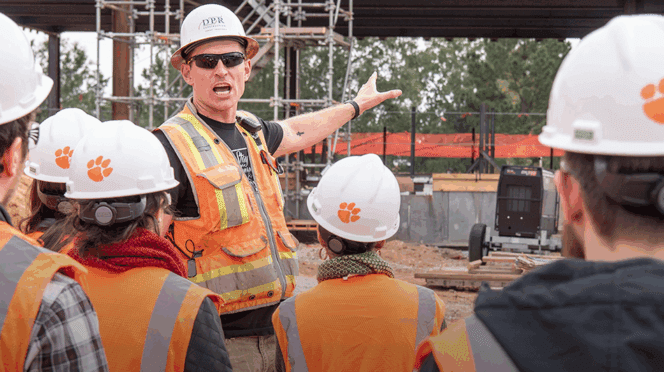 A DPR representative is seen pointing to the construction behind him as a group of students in white hardhats with an orange Tiger Paw are seen in the foreground.