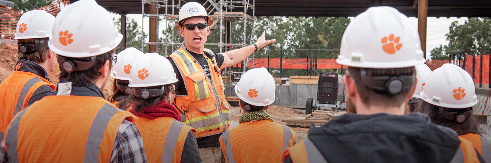 A DPR representative is seen pointing to the construction behind him as a group of students in white hardhats with an orange Tiger Paw are seen in the foreground.