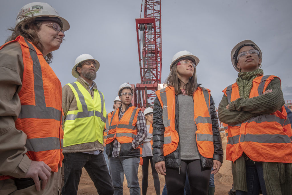 A professor and 4 students stand looking up at the building under construction. A large red crane is seen in the background.