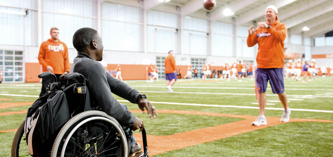 From his wheelchair, Marsden Miller throws a football with a man clad in purple and orange gear on the football field inside the Clemson indoor practice facility.