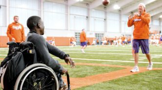 From his wheelchair, Marsden Miller throws a football with a man clad in purple and orange gear on the football field inside the Clemson indoor practice facility.