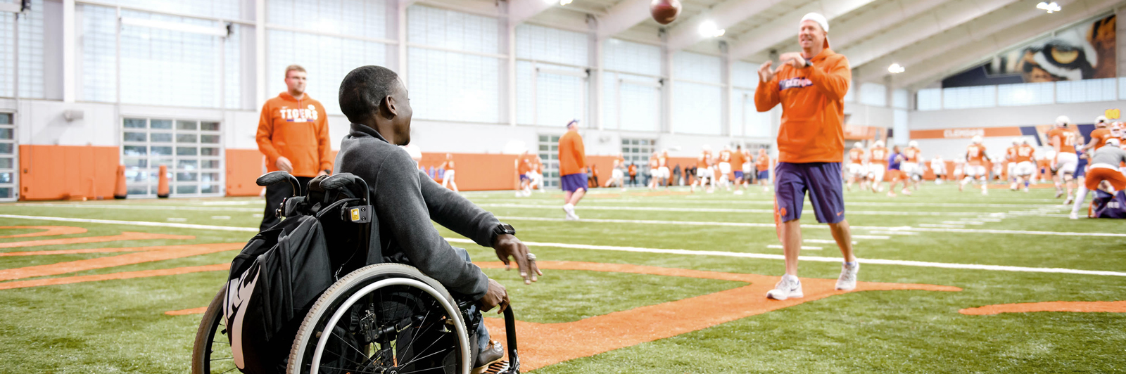 From his wheelchair, Marsden Miller throws a football with a man clad in purple and orange gear on the football field inside the Clemson indoor practice facility.