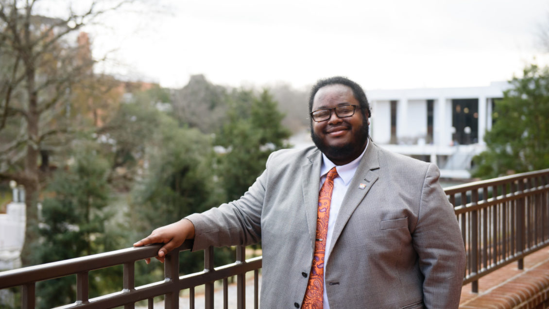 Jerad Green stands with his hand leaning on a metal rail. The Clemson Library is seen in the background.