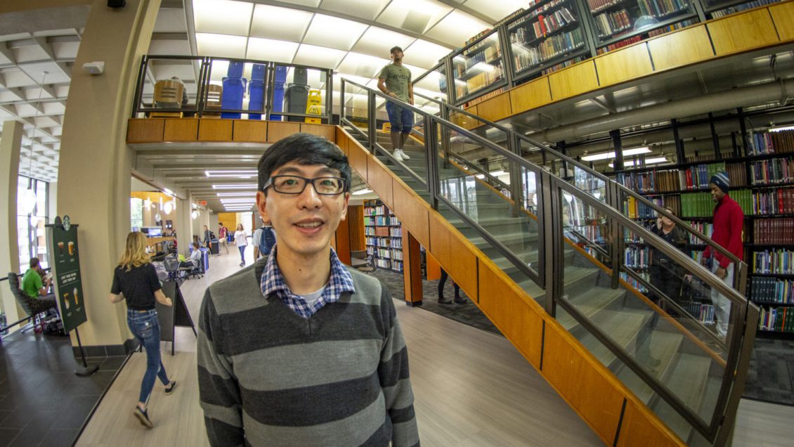 A man with a striped sweater and glasses stands in the library, with book cases and a stair case next to him and students walking around behind him.