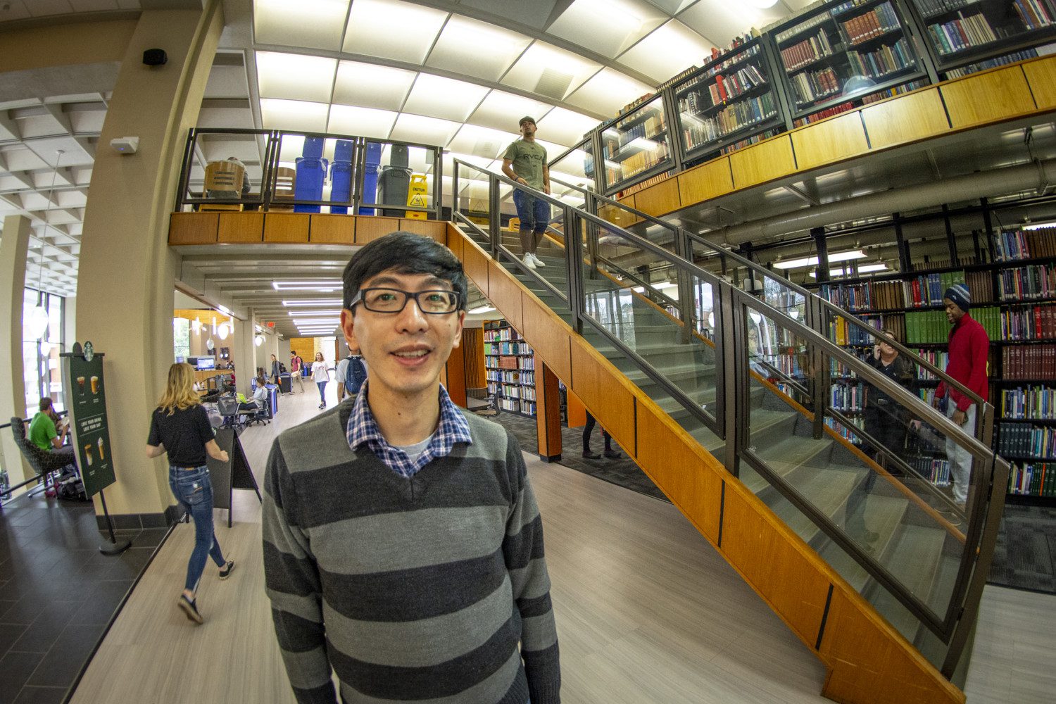 A man with a striped sweater and glasses stands in the library, with book cases and a stair case next to him and students walking around behind him.