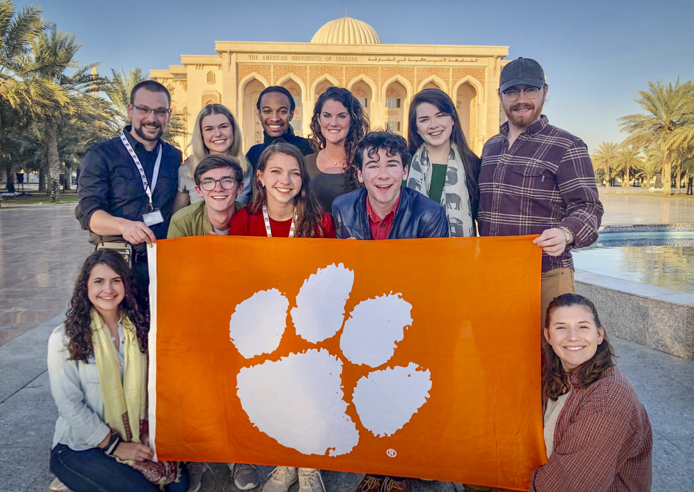Ten people stand in a group holding a large Clemson Tiger paw flag, with a Middle Eastern-style building and palm trees behind them