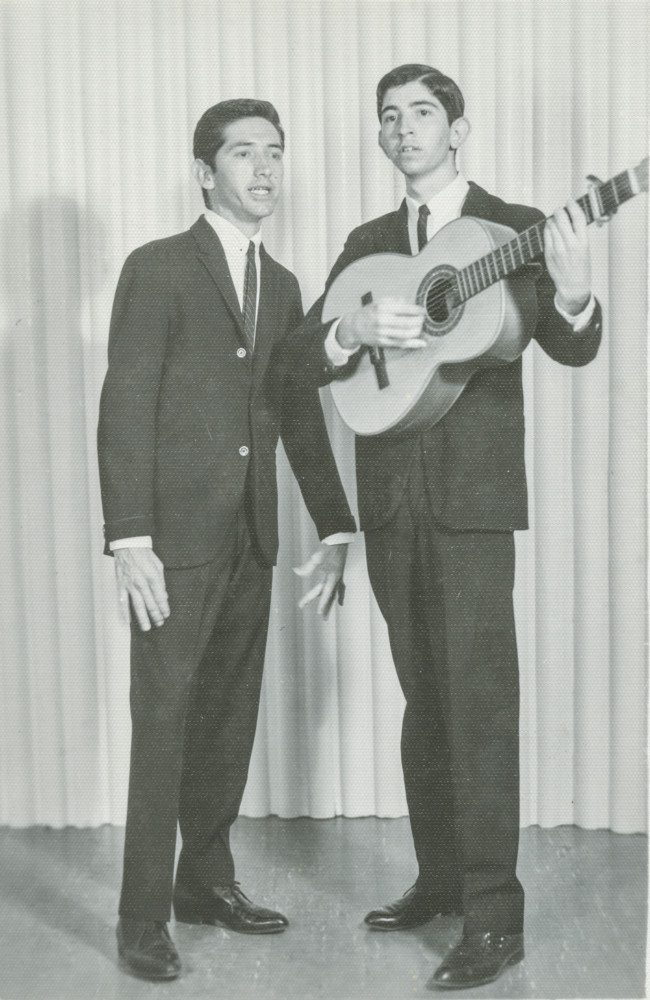 Two men in suits stand in front of a show curtain singing. One man is playing guitar
