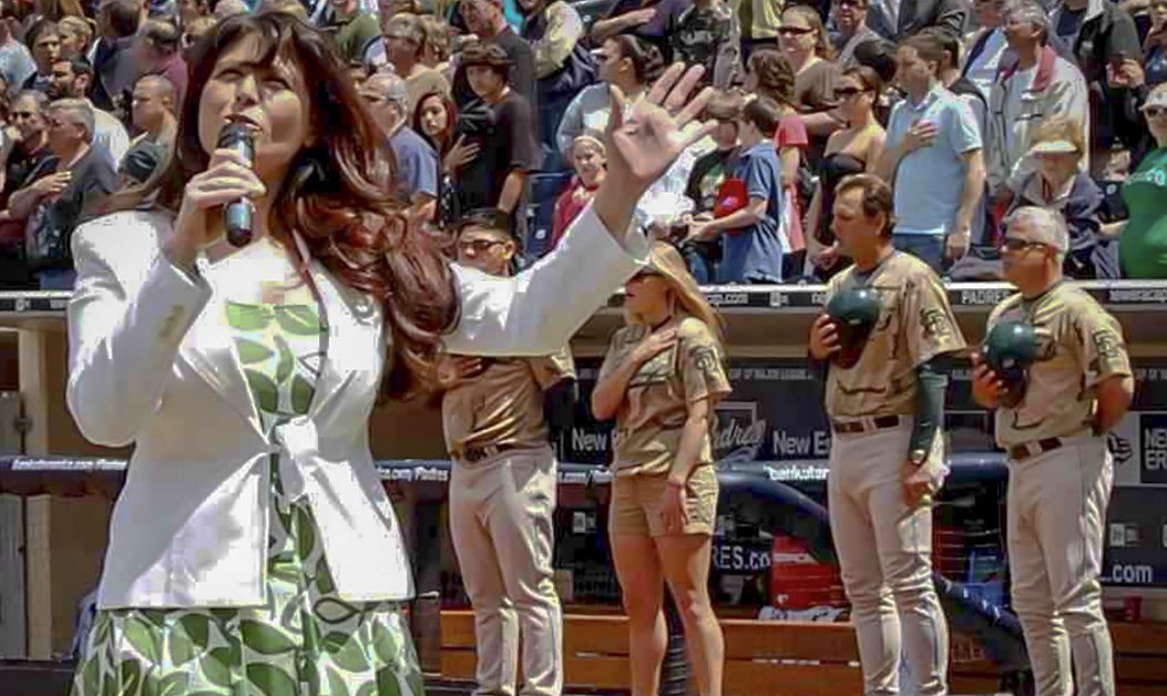 A woman in a flowery dress and white jacket sings into a microphone with baseball players and a crowd-filled stand behind her.