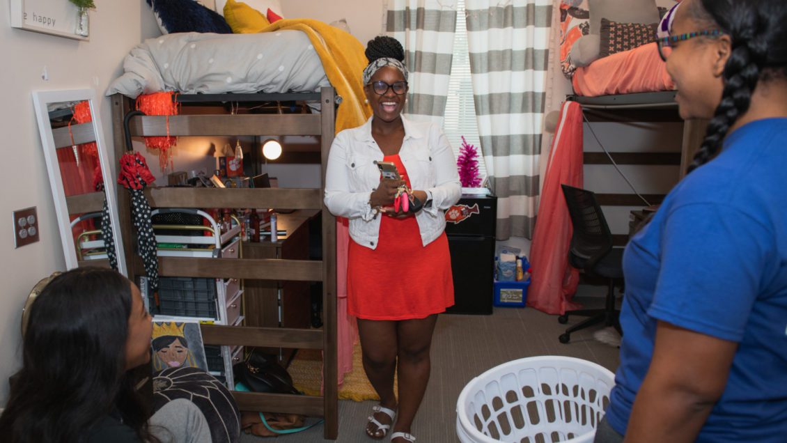 Three women stand in a dorm room laughing.