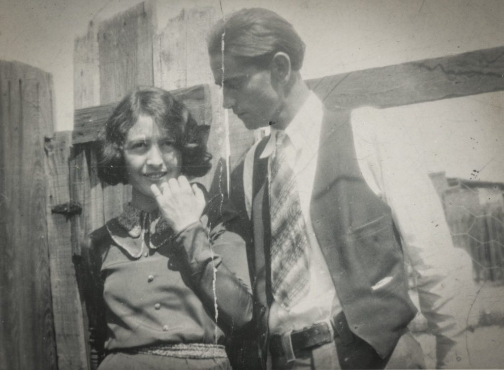 Black and white photo of a man and woman, dressed up and standing in front of a wooden fence.