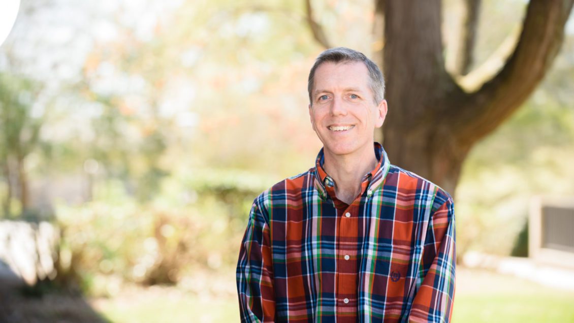 A man in a plaid shirt stands in front of a tree for a photo.