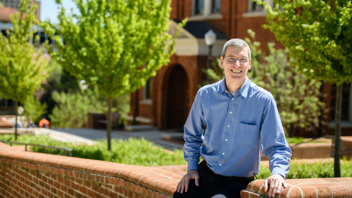 David Taylor leans against a short, red-brick wall with Hardin Hall in the background