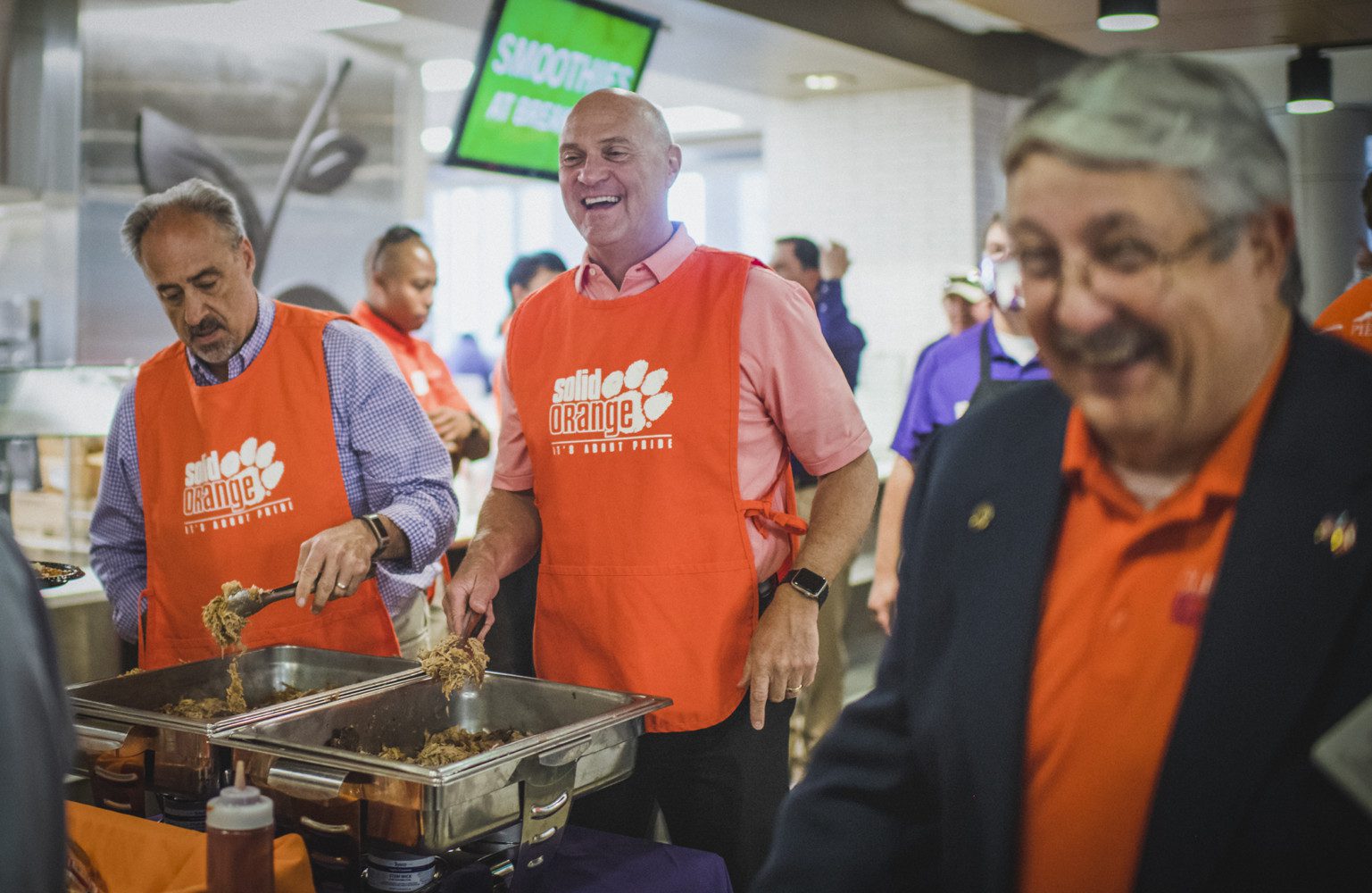 President Clements, in an orange Clemson apron, serves pulled pork out of a large stainless steel conatiner