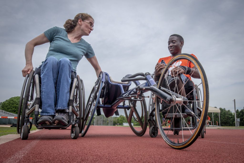 Two people in wheelchairs sit on either side of a racing wheelchair, with a cloudy sky behind them.