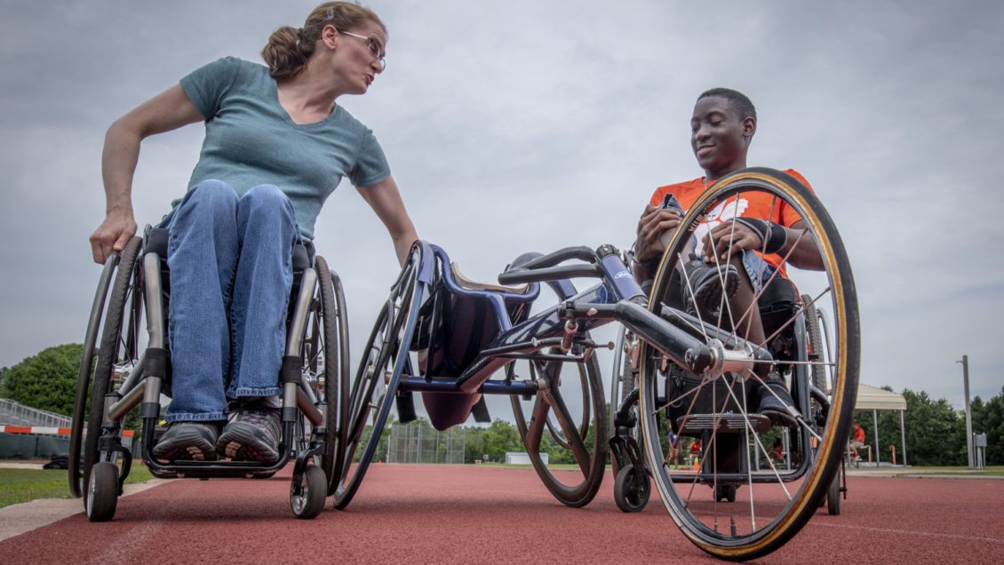 Two people in wheelchairs sit on either side of a racing wheelchair, with a cloudy sky behind them.