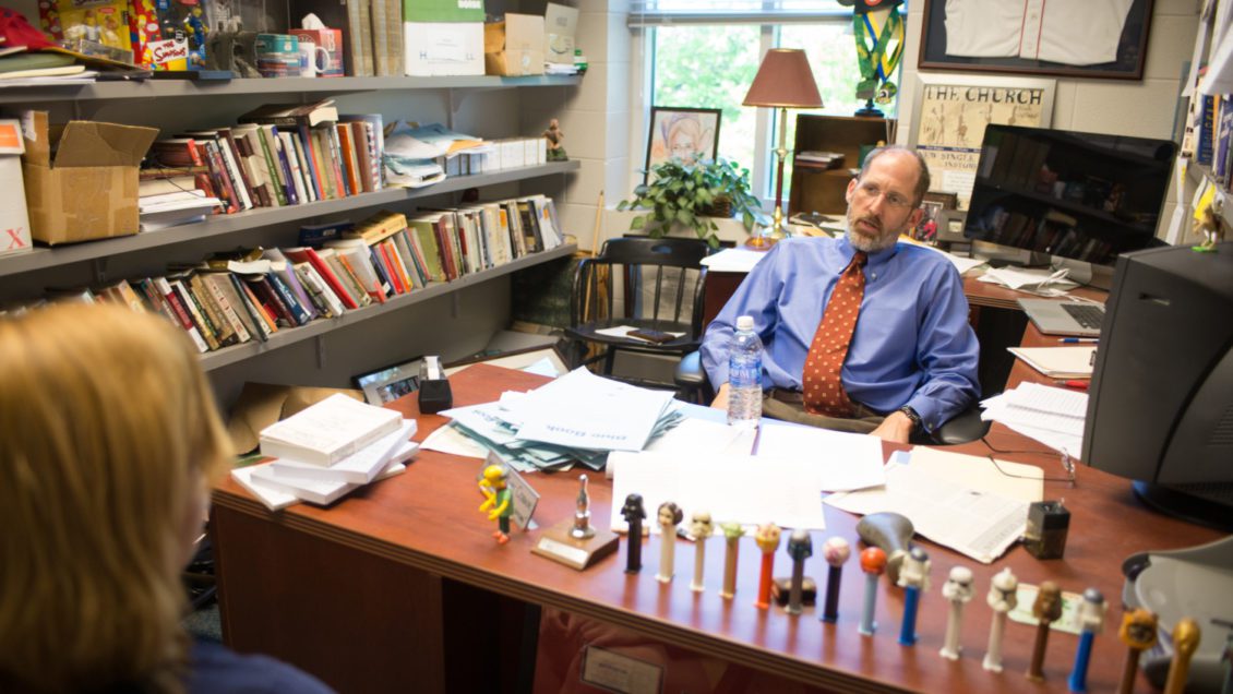 Professor casually sits behind the cluttered desk in his office, chatting with a student.
