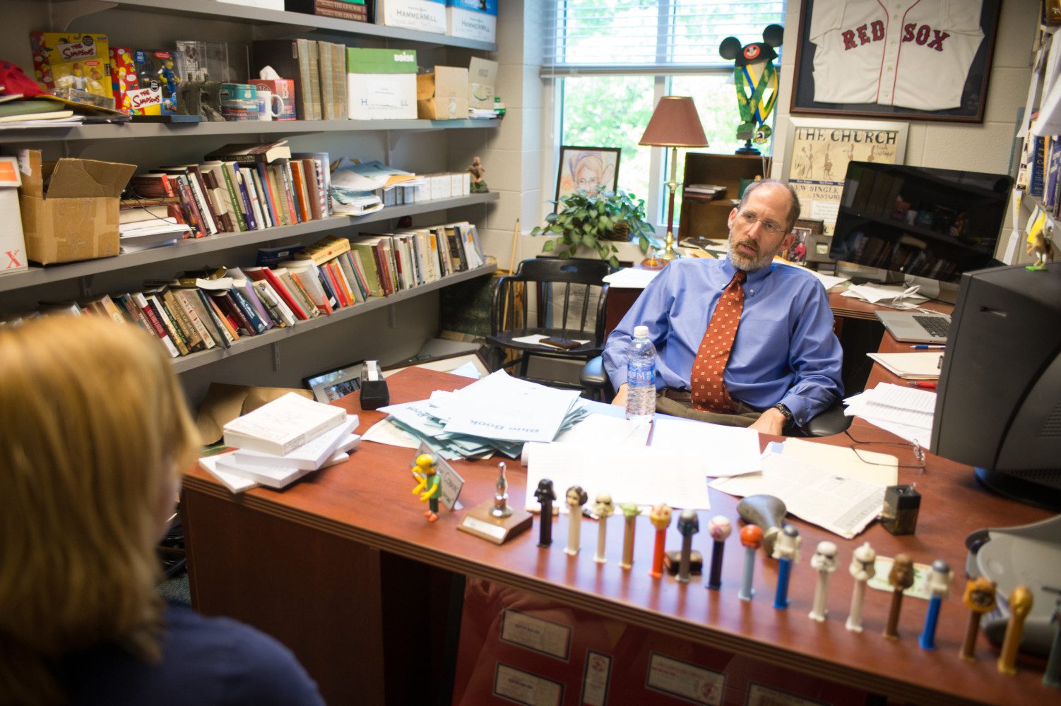 Professor casually sits behind the cluttered desk in his office, chatting with a student.