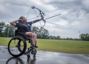 A woman in a wheelchair raises a bow and arrow in front of a cloudy sky.