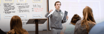 A male student teacher stands at the front of a classroom holding papers as he leads his class through a lesson.