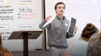 A male student teacher stands at the front of a classroom holding papers as he leads his class through a lesson.