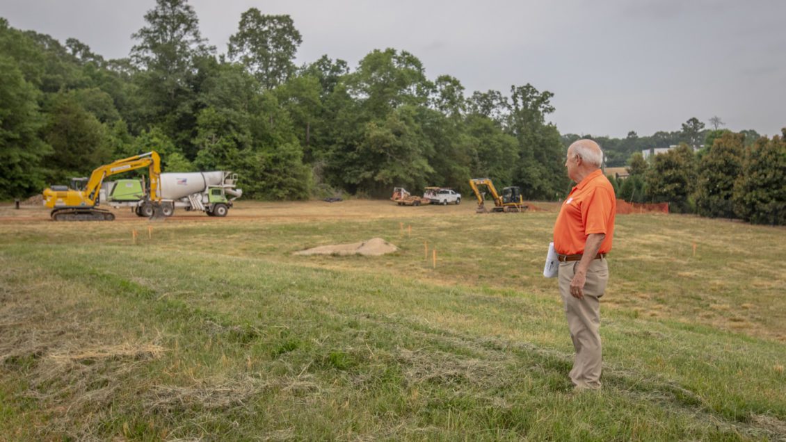 A man in an orange shirt looks over a grass and dirt field with construction equipment in the distance