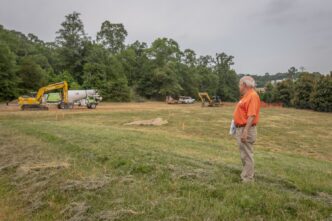 A man in an orange shirt looks over a grass and dirt field with construction equipment in the distance