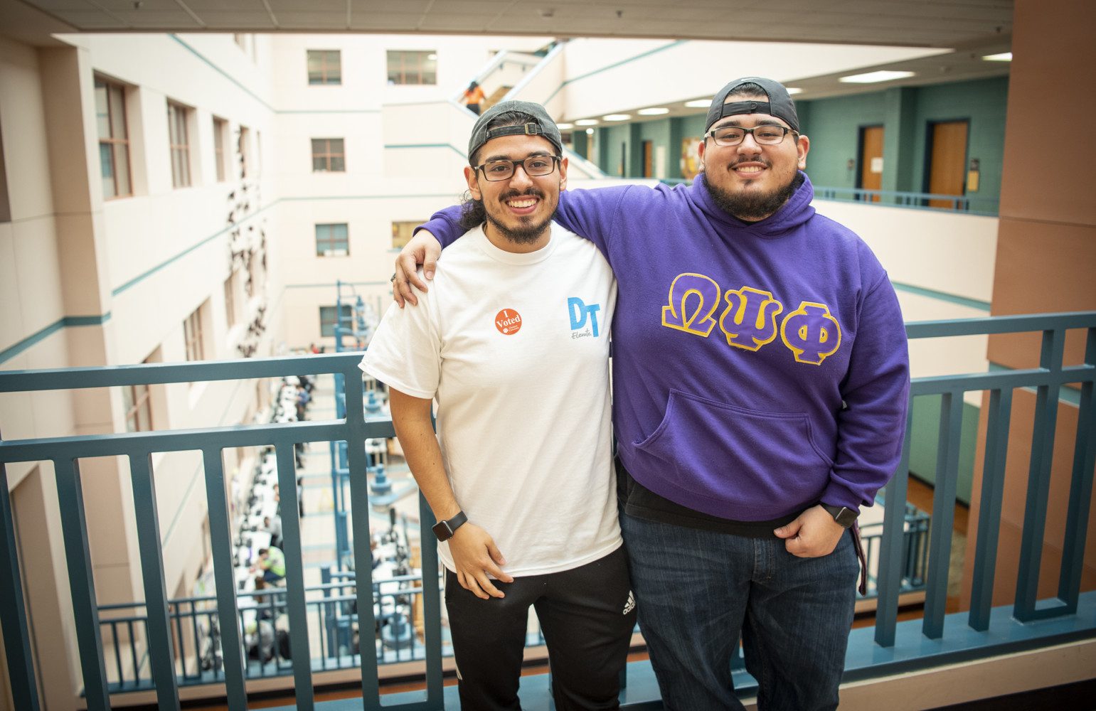 Two Hispanic-American brothers, each wearing glasses and backward baseball caps, pose with their arms around each other