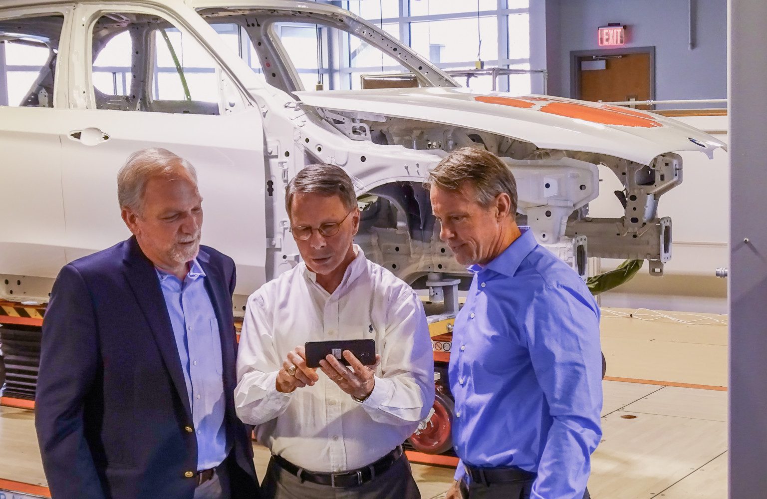 Three men look at a tablet while standing in front of a vehicle that's in the process of being assembled.