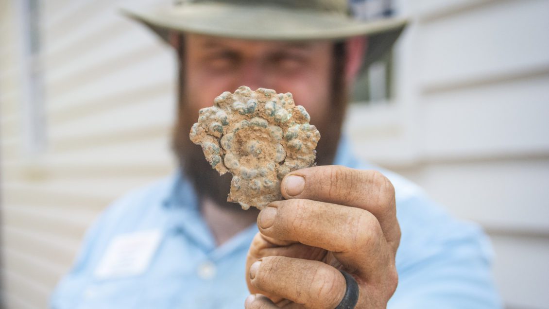 A man holds up a decorative round piece of brass, with dirt all over it and his hands