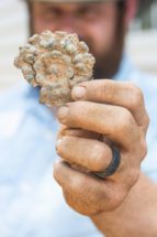 An out-of-focus man holds up a decorative round piece of brass with dirt all over it and his hand