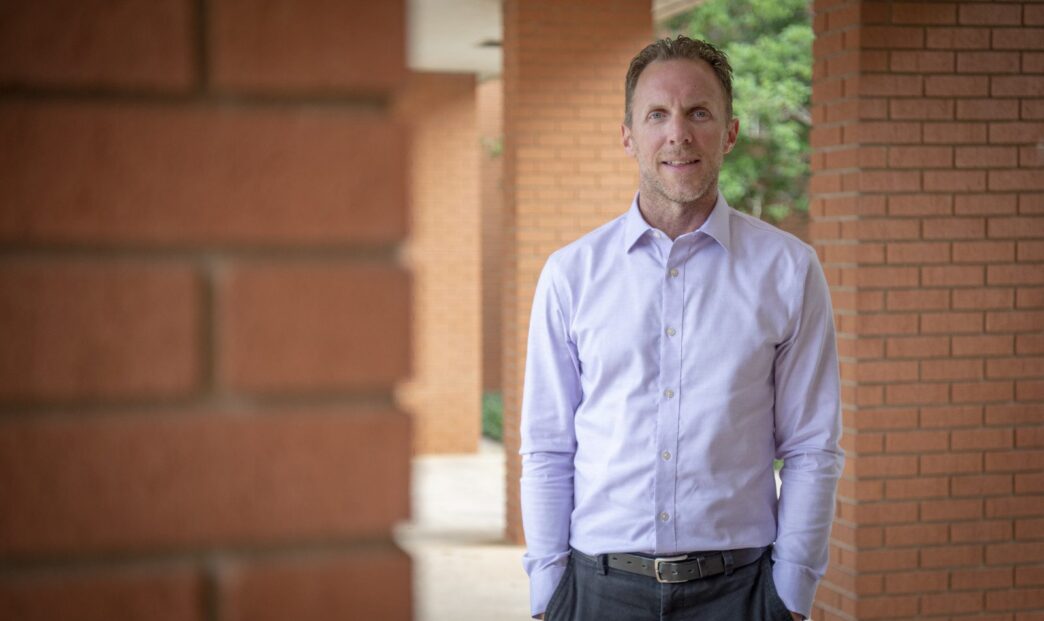 A man wearing a violet-white button-up shirt stands in front of green foliage