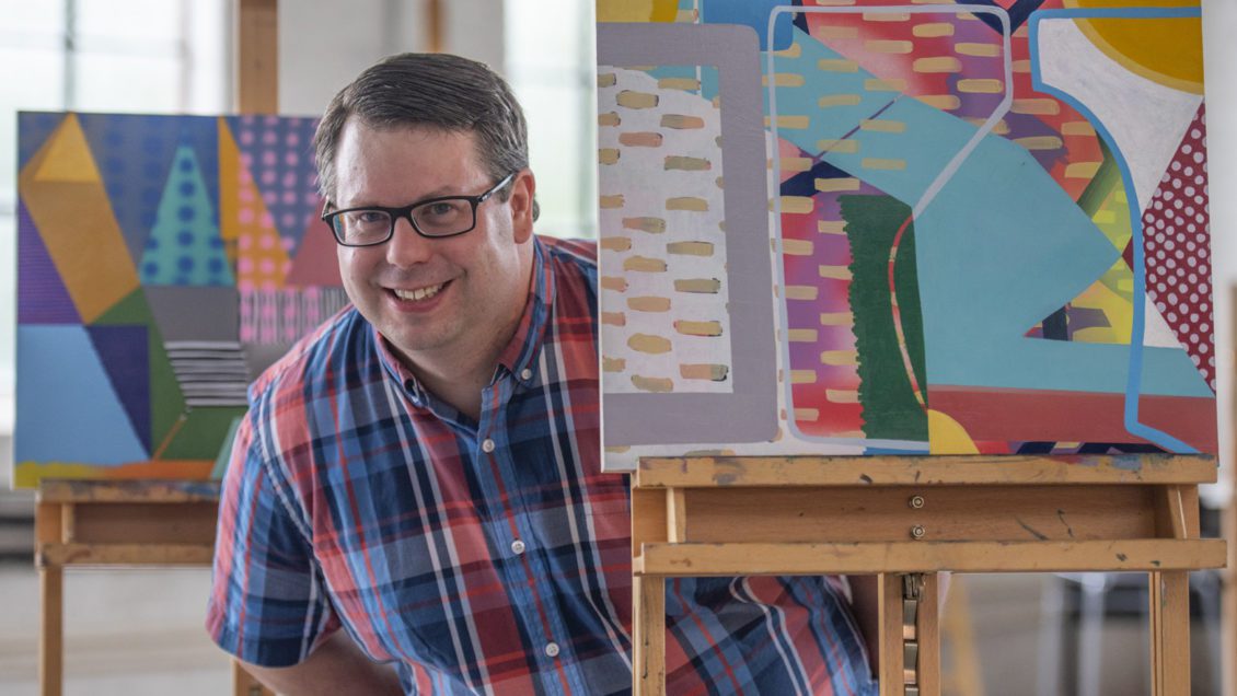 A man wearing a plaid shirt stands next to three easels holding colorful abstract paintings