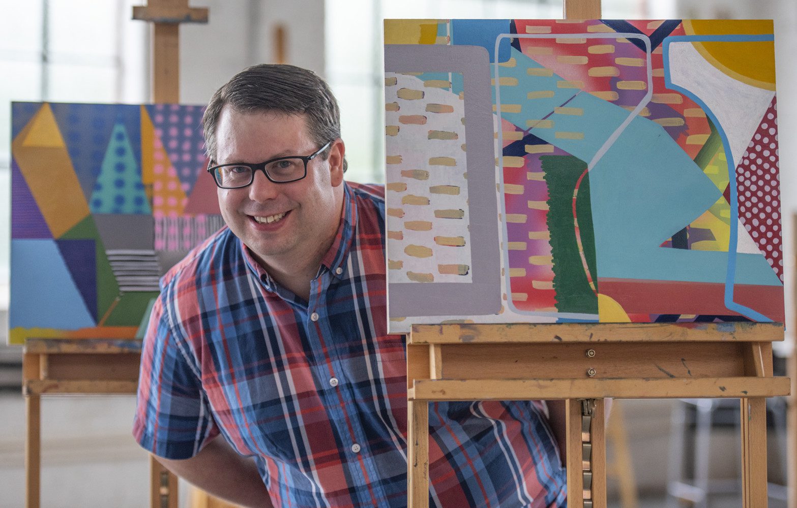 A man wearing a plaid shirt stands next to three easels holding colorful abstract paintings