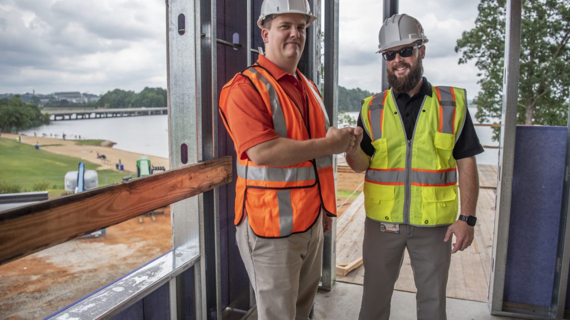Two men in hard hats and safety vests fist-bump in front of an open window.