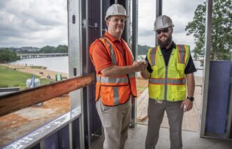Two men in hard hats and safety vests fist-bump in front of an open window.