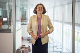 A woman in a cream-colored jacket and black skirt holds the edges of the jacket and laughs with a large atrium with wall-to-ceiling windows behind her