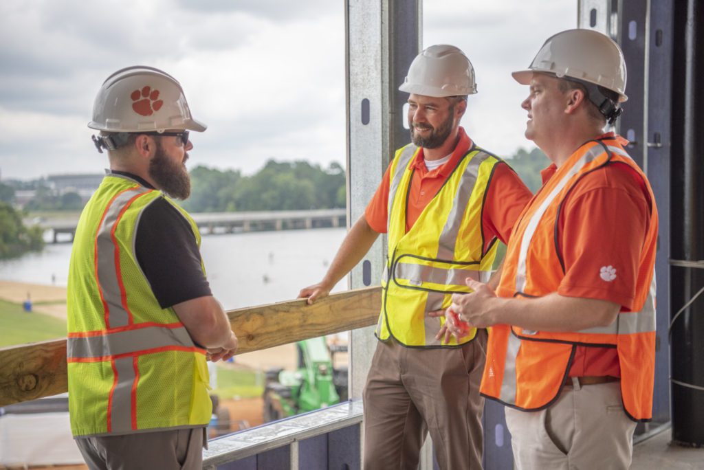 Three men wearing hard hats and safety vests chat in front of an open window, through which Lake Hartwell can be seen in the background.