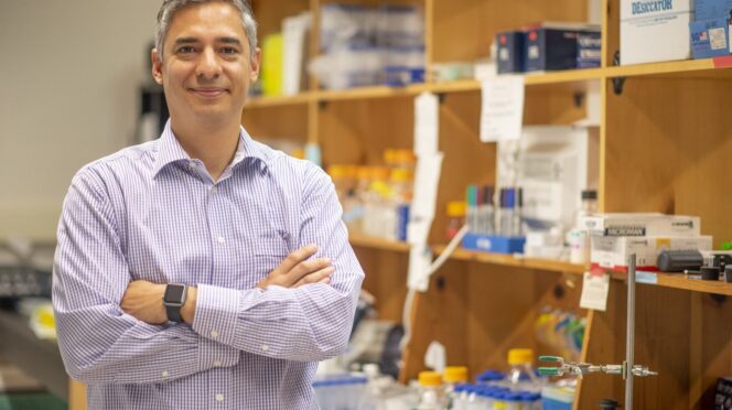 A man stands with arms folded, in front of shelves containing laboratory equipment.
