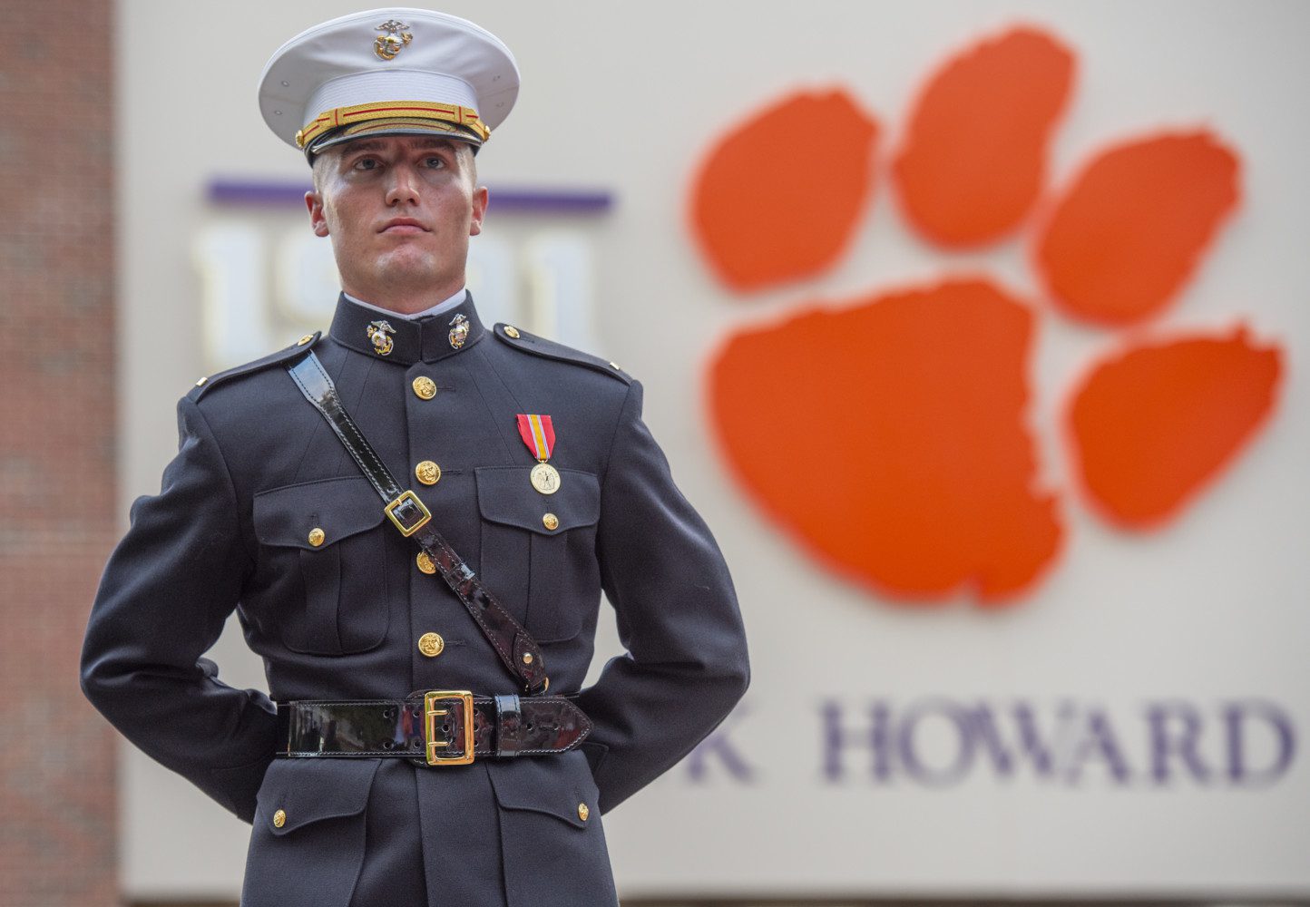 A Marine in dress uniform stands with arms behind his back, a large Clemson Tiger paw attached to Memorial Stadium behind him, over his shoulder